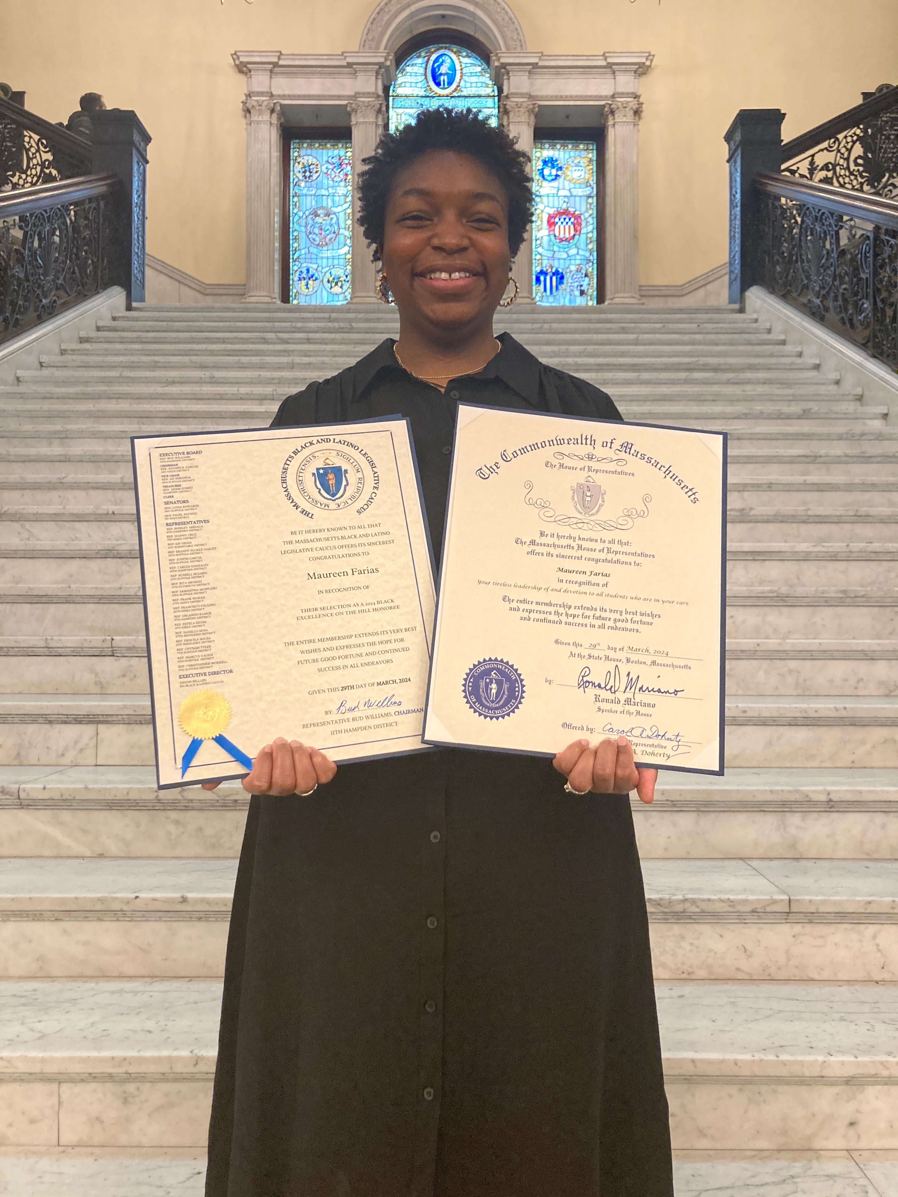 Maureen Farias holding up her citations and recognition in front of steps at the State House